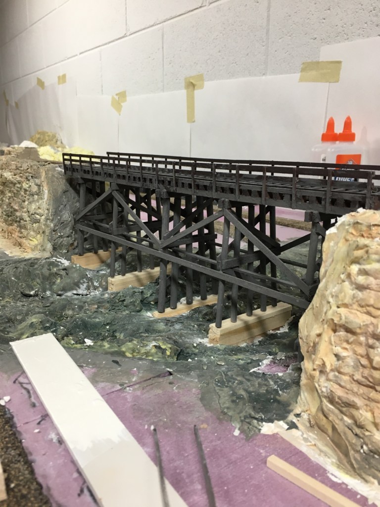 A large scratch-built trestle is in place over the river that will flow beneath it.  Poster board is on the wall where backdrop scenes will be painted.