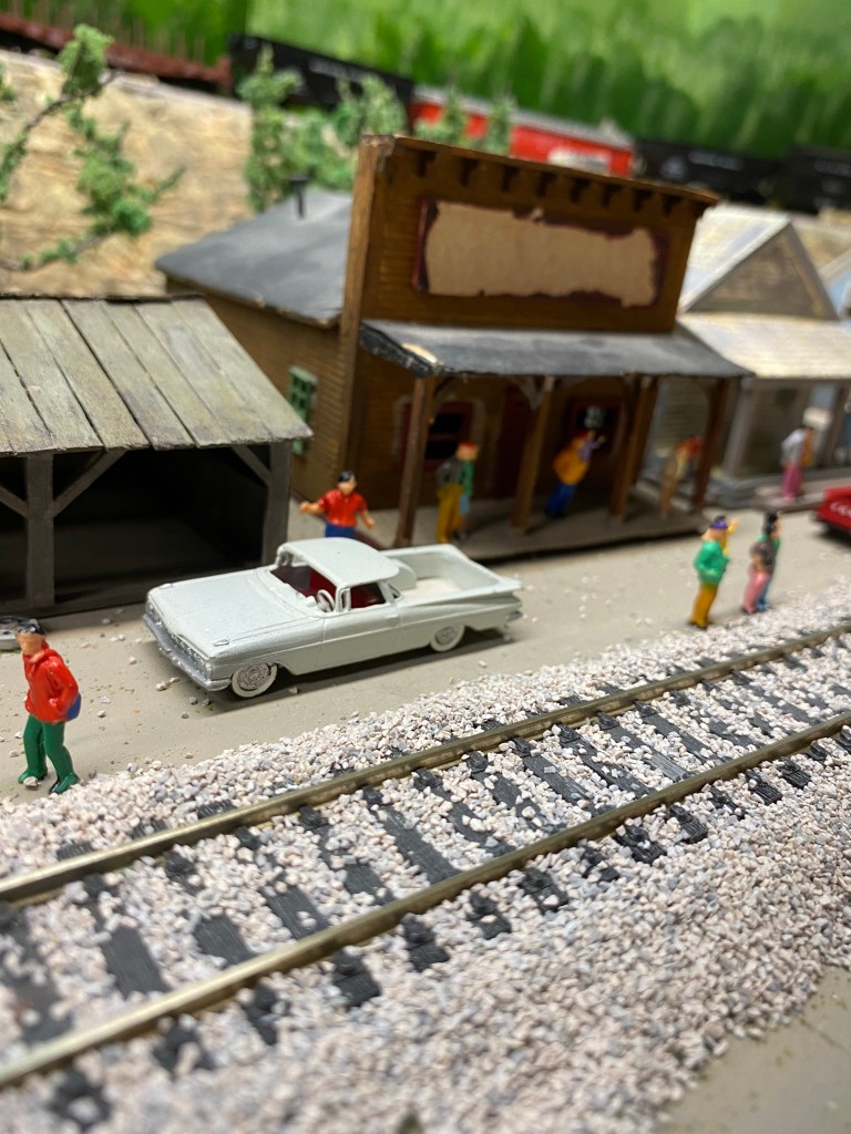 Shops along the tracks with people and cars near track.