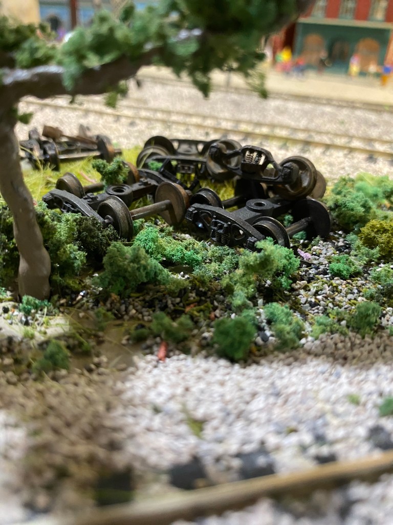 Several old rail car trucks sit abandoned under a tree, surrounded by weeds.