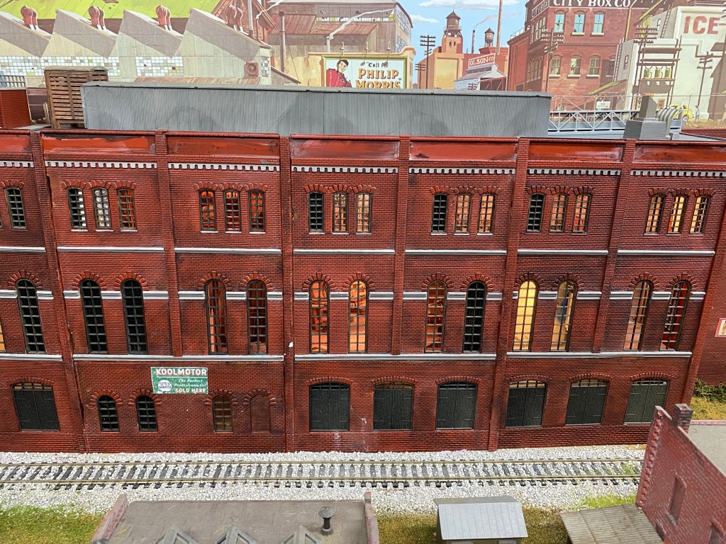 A distant view of Maple Valley Manufacturing with buildings in the foreground, the supply track, and the backdrop photo of other factories.