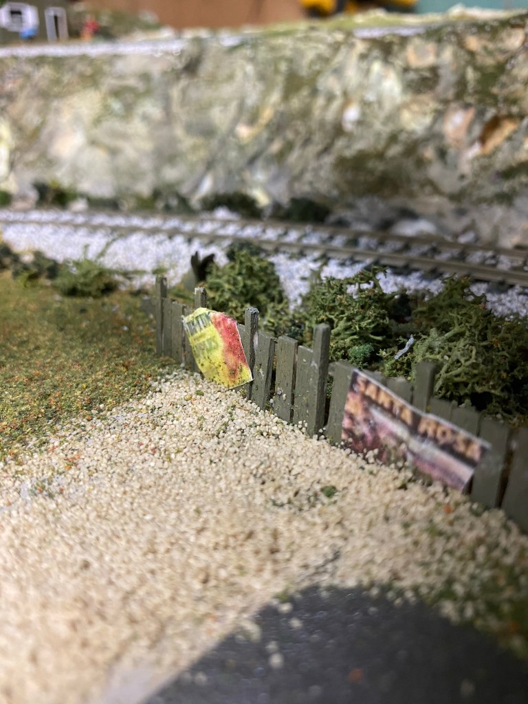 The Maple Valley curve, with a poster-covered fence, bushes, a road, and rocks.