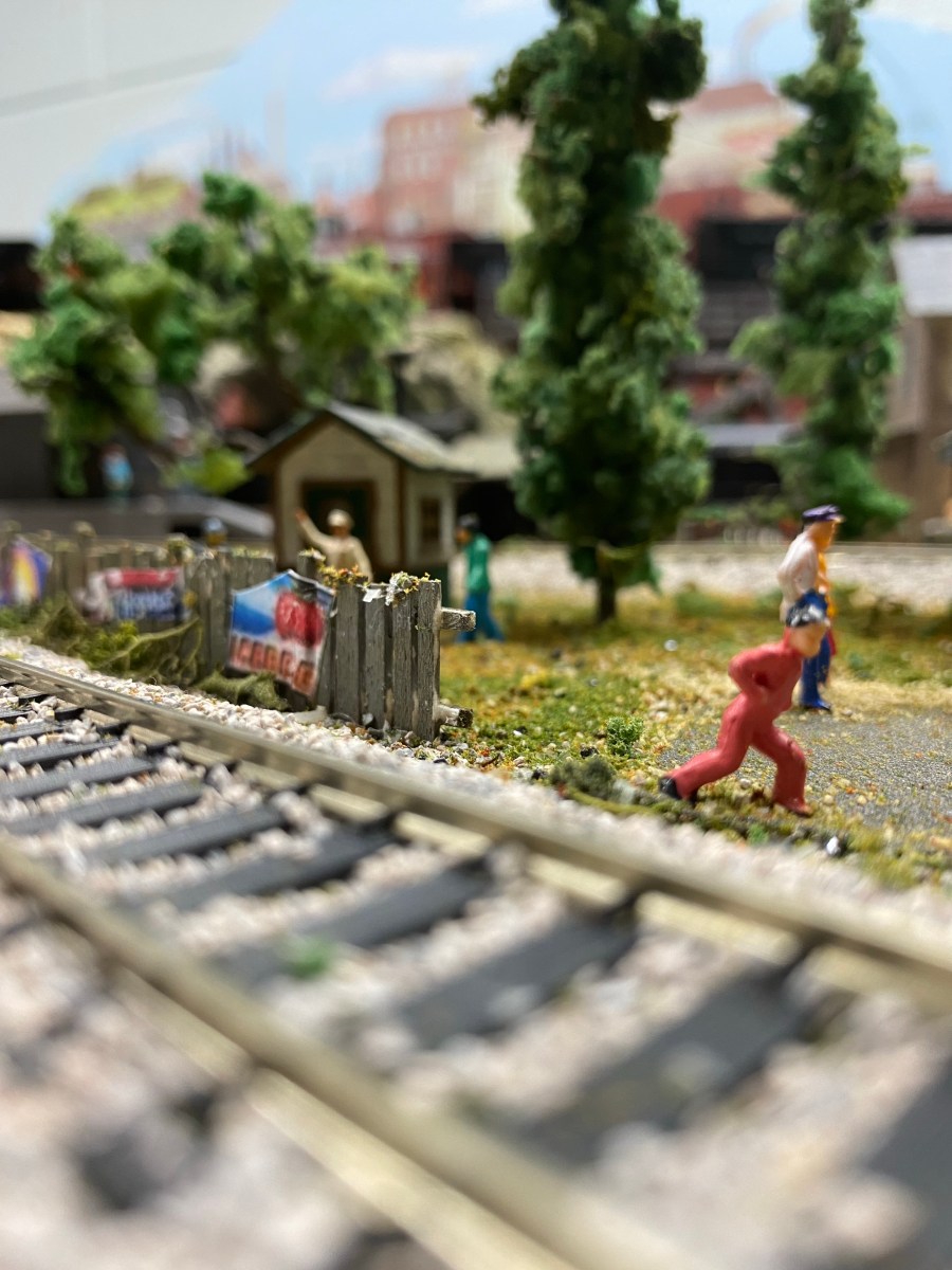 A train track in the foreground, with a fence with posters, several people, trees, and a shed.