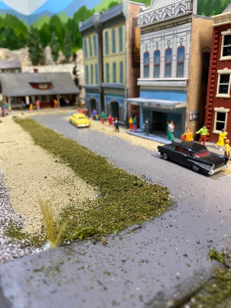 Two cars sitting on a street, buildings, ground cover, weeds