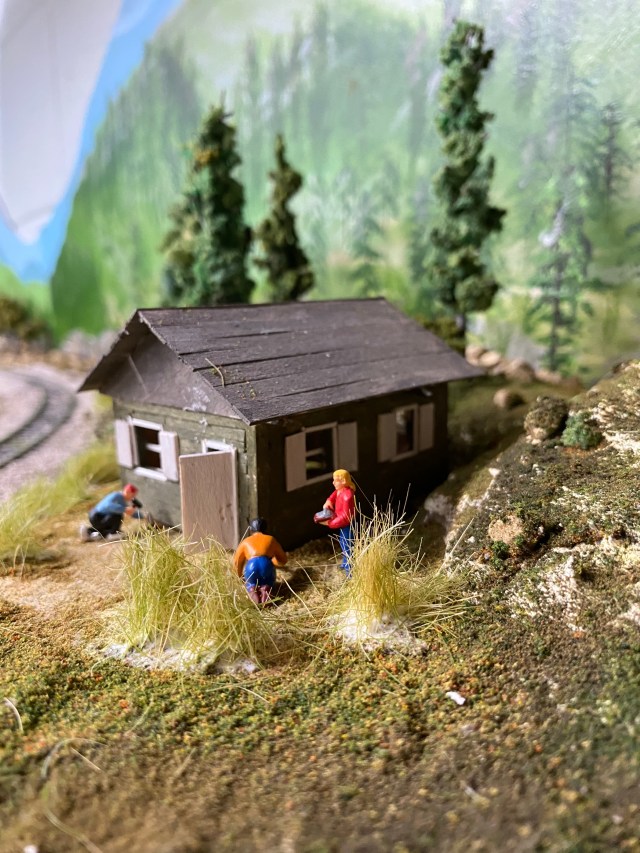 Small cabin, people working, trees, weeds, rocks, grass, backdrop