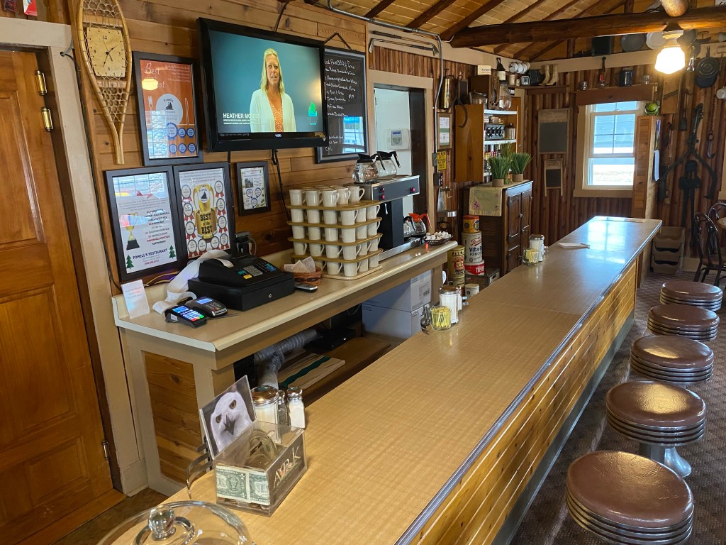 Vintage snack bar and stools, stacked coffee mugs, coffee pot, log wall, TV, ceiling, antique decor