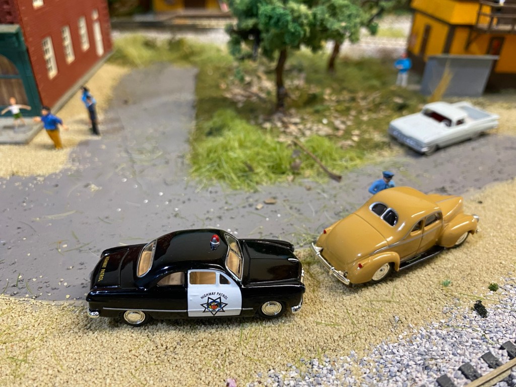 Police car, officer, two cars, people, trees, grass, buildings